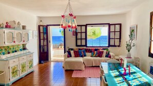 A spacious living room with a large sofa, a dining table, and a view of the sea through a window. - Spacious Beachfront Villa (Porto Columbu-Perd'È Sali)