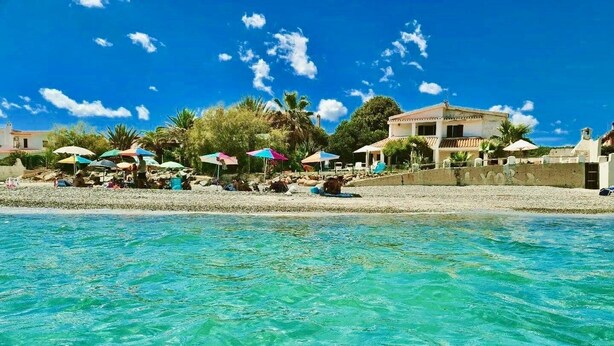 A beach with clear turquoise water, umbrellas, and buildings in the background. - Spacious Beachfront Villa (Porto Columbu-Perd'È Sali)