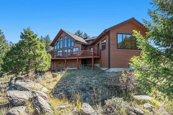 A two-story log cabin with a balcony, surrounded by trees and rocks.