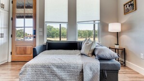 A bedroom with a large bed, a door with glass panels, two windows with blinds, a bedside table with a lamp, and a framed picture on the wall.
