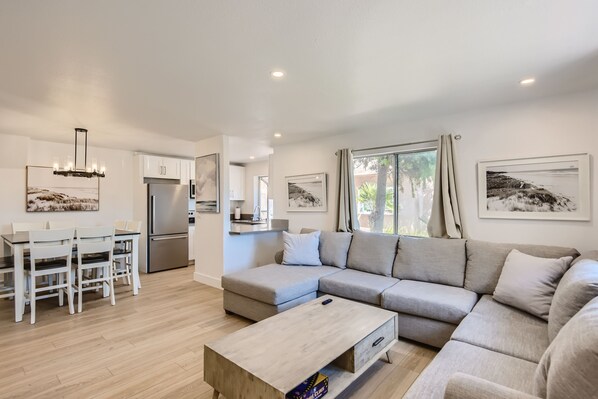 A modern living room with a sectional sofa, a dining table with chairs, a kitchen area, and artwork on the walls.