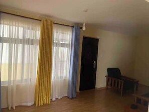Um quarto com grandes janelas cobertas por cortinas finas brancas e amarelas, piso de madeira, uma porta escura e um pequeno banco de madeira.