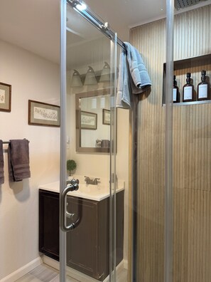 A modern bathroom with a glass shower enclosure, a vanity with a dark cabinet, and a shelf with toiletries.