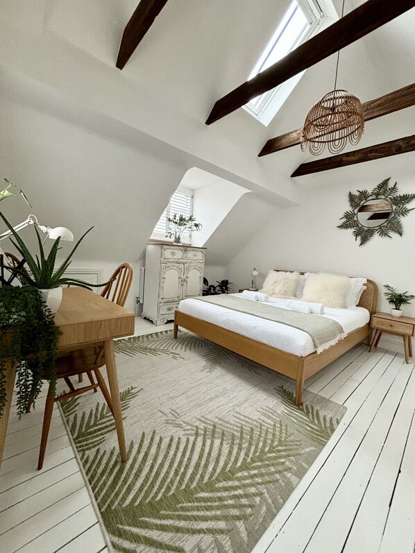 A bedroom with a sloped ceiling, a skylight, a bed with white bedding, a wooden chair, a small table, and a potted plant.