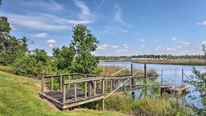 Un muelle de madera que se adentra en un lago tranquilo, rodeado de abundante vegetación y un cielo despejado.