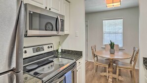Una cocina con armarios blancos, un refrigerador de acero inoxidable, un microondas integrado y una estufa. Hay una mesa de comedor con sillas y una planta en maceta.