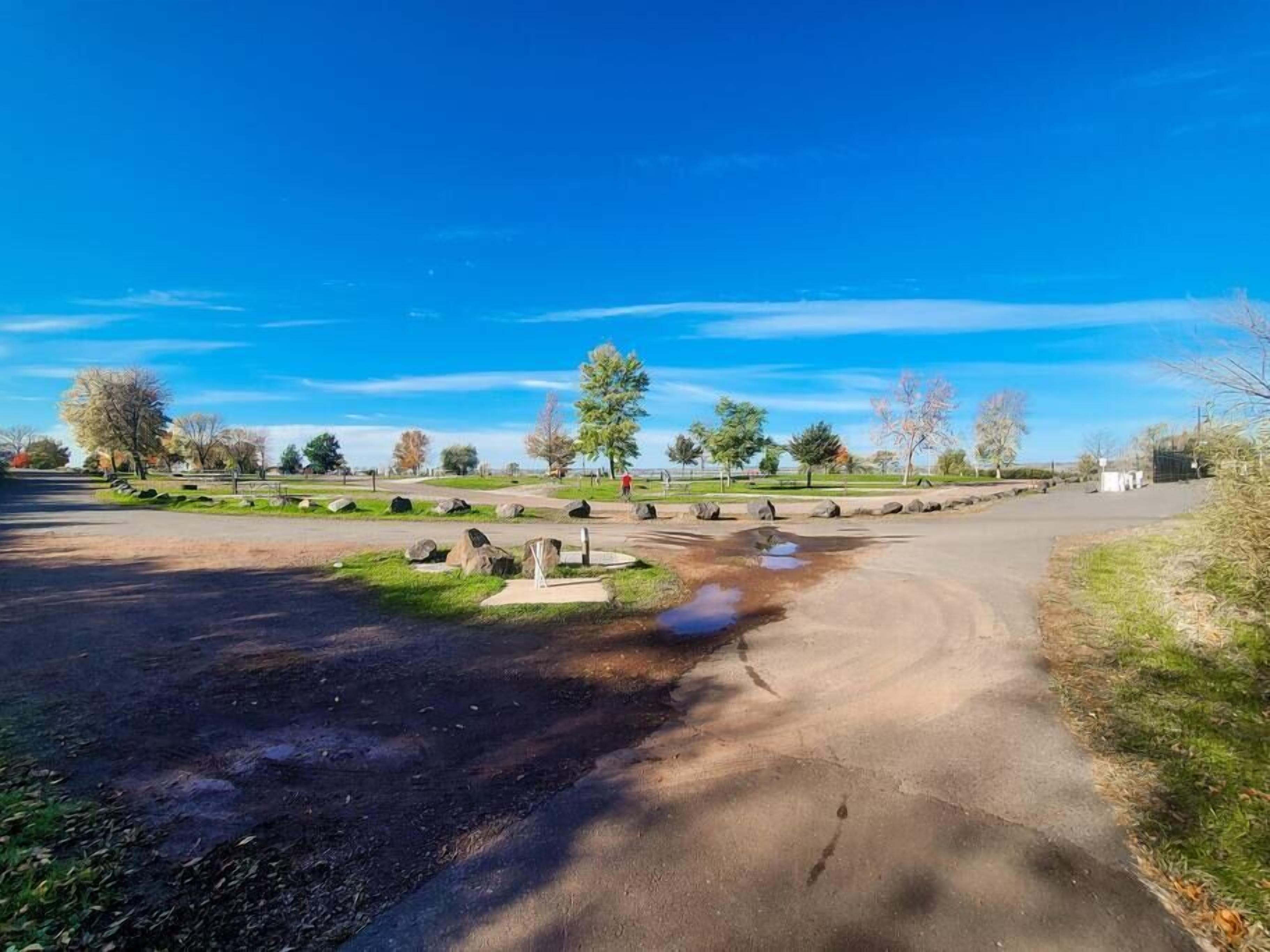A paved parking area with a grassy median, trees, and a clear blue sky.