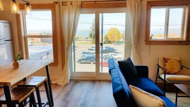 A modern living room with a blue sofa, wooden dining table, and a view of a parking lot through large windows.