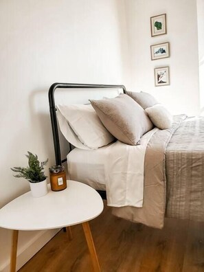 En pænt redt seng med en beige dyne og puder, et lille hvidt bord med en plante og et lys, og tre billeder hængende på væggen.