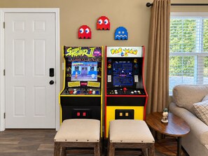 Zwei Spielautomaten, einer für Street Fighter II und einer für Pac-Man, in einem Raum mit einer Tür, einem Fenster mit Jalousien und einem Sofa.