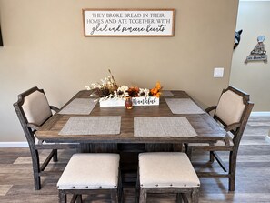 A dining table with a wooden finish, surrounded by chairs with cushioned seats. A decorative sign is mounted on the wall above the table.