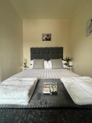 A neatly made bed with a grey headboard, white linens, and a small bedside table with a lamp and a plant.