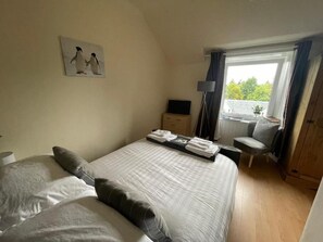 A bedroom with a bed, pillows, a bedside table, a lamp, a chair, a window with curtains, and a painting on the wall.