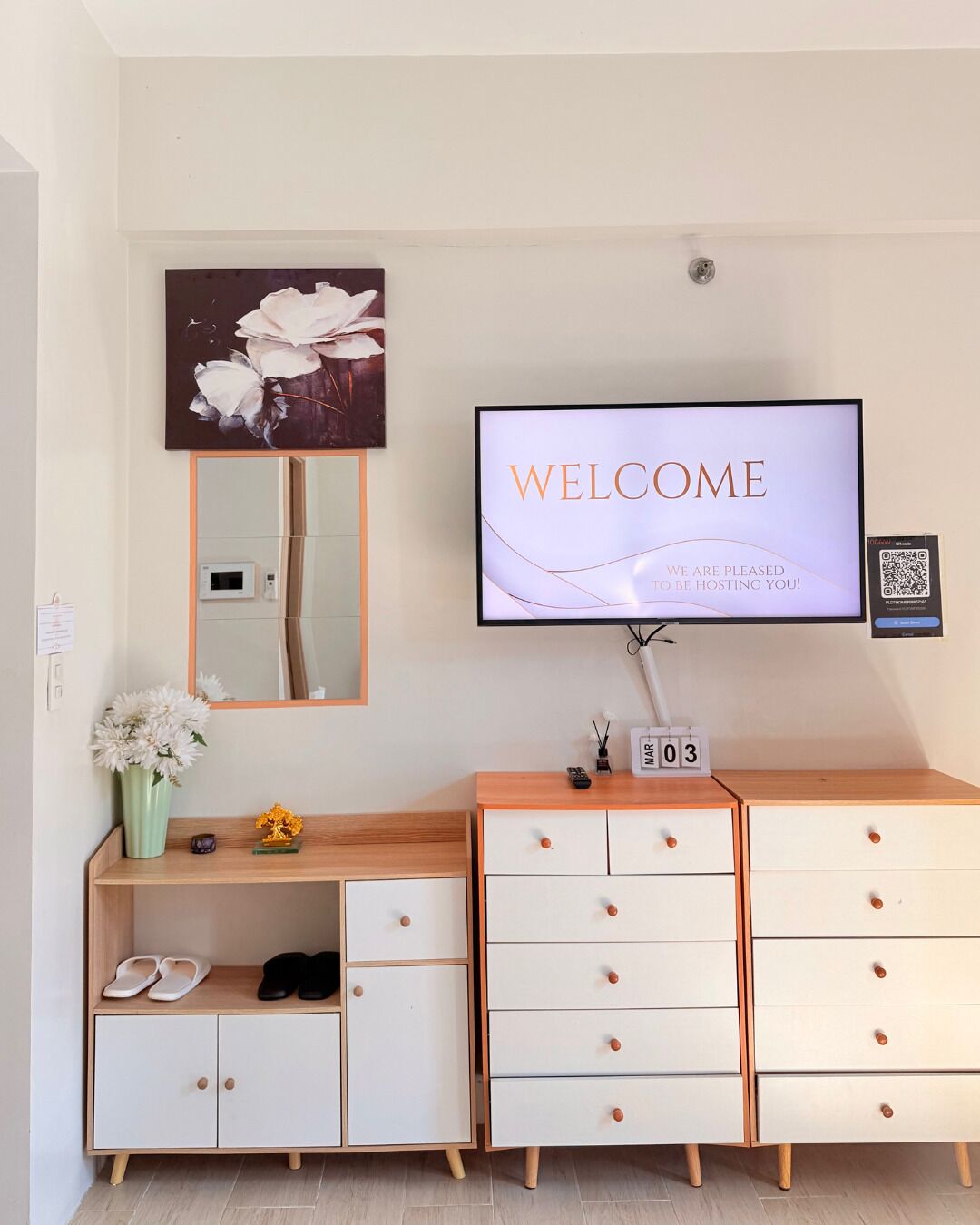 Een moderne hotelkamer met een grote flatscreen-tv waarop een welkomstbericht staat, een houten dressoir met lades, een spiegel en een vaas met bloemen op het dressoir.