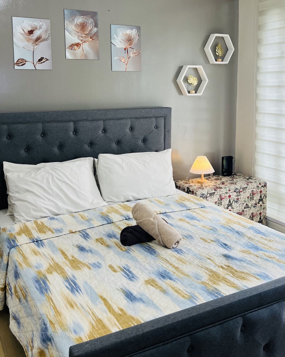 Un dormitorio con cabecera capitonada, cama con colcha estampada, almohadas blancas, una mesita de noche con lámpara y decoración en la pared.
