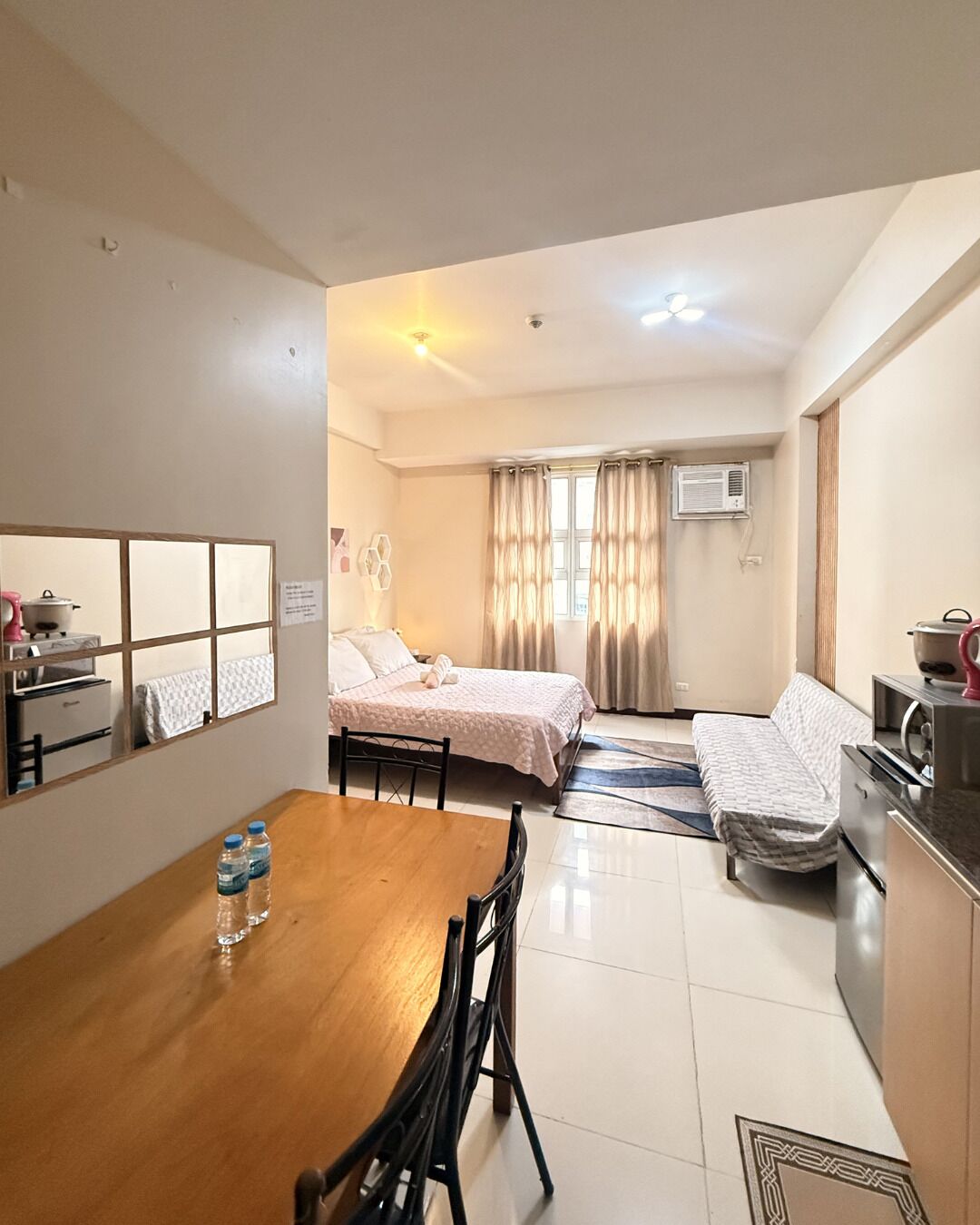 A compact hotel room with a dining table, two beds, a kitchenette, and a large window with curtains.