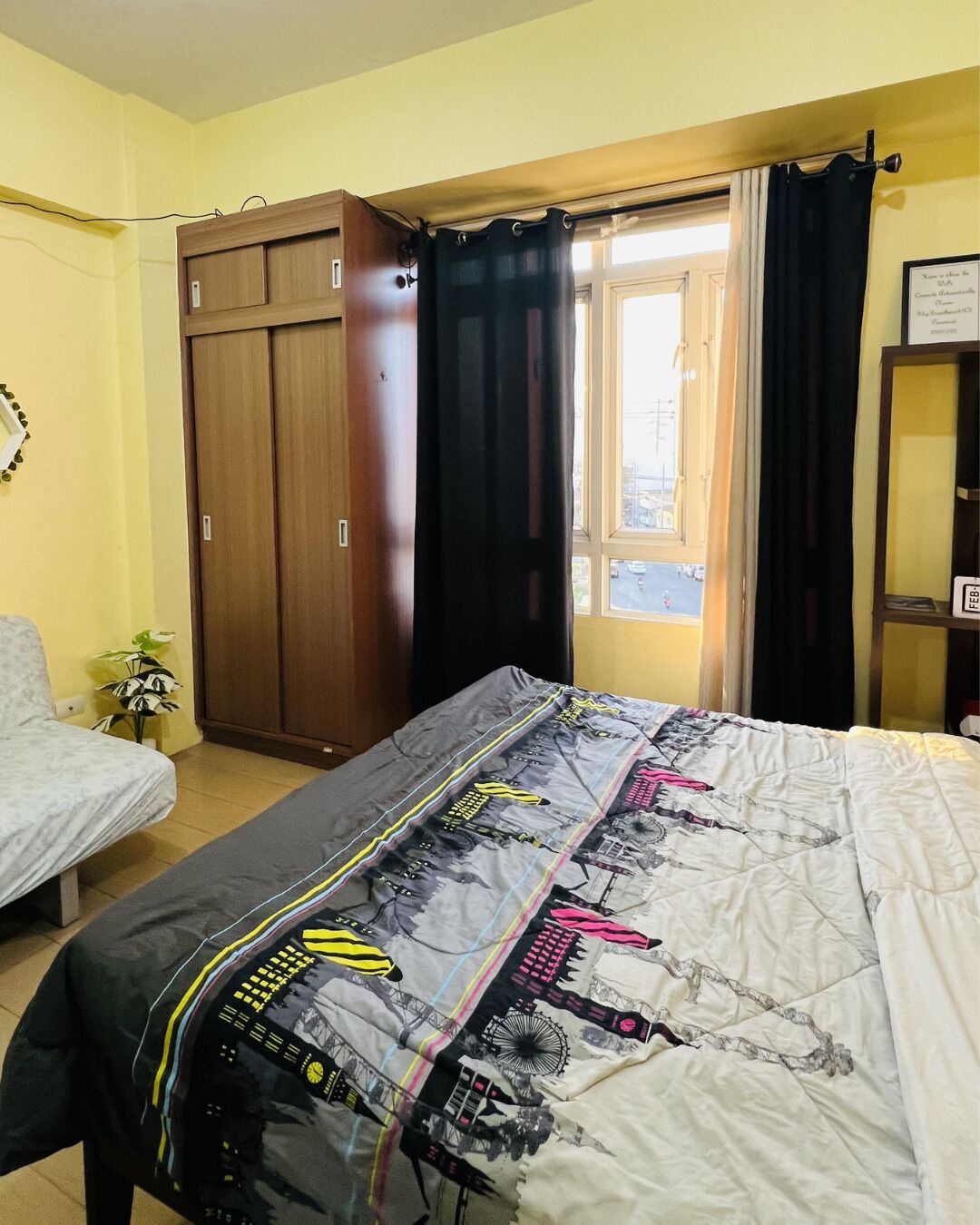 Un dormitorio con cama, armario, ventana con cortinas y una planta pequeña.