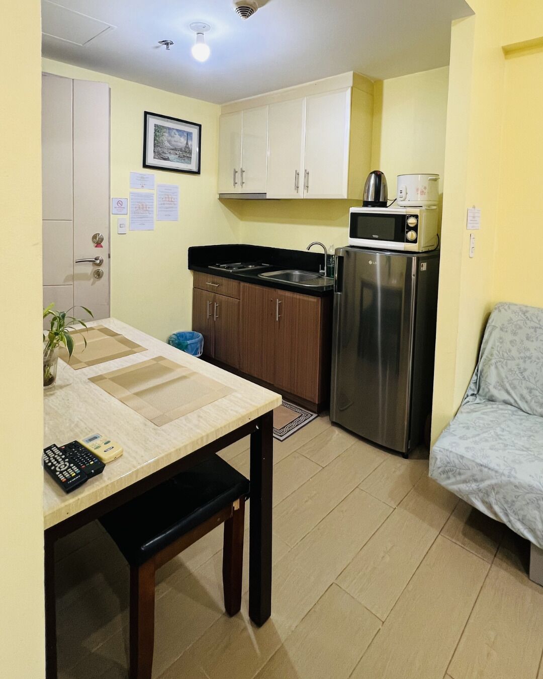 A compact kitchen with a refrigerator, microwave, and sink. There is a dining table with chairs and a small plant.