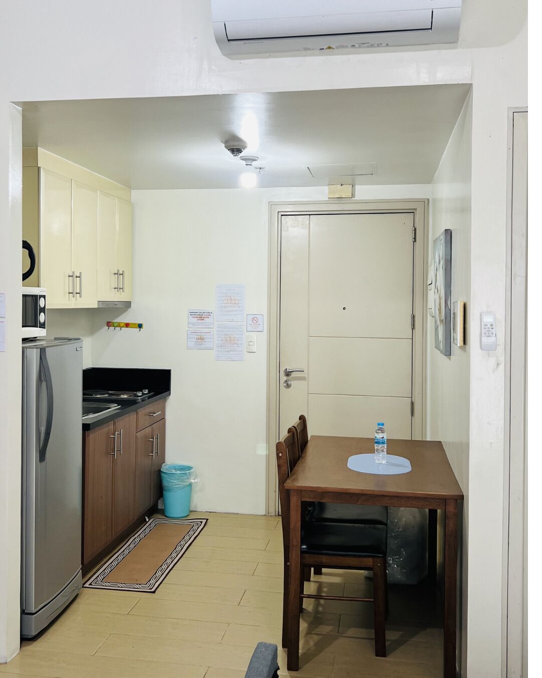 A compact kitchen with a refrigerator, microwave, and stove, a dining table with chairs, a blue trash bin, and a door with a light switch.
