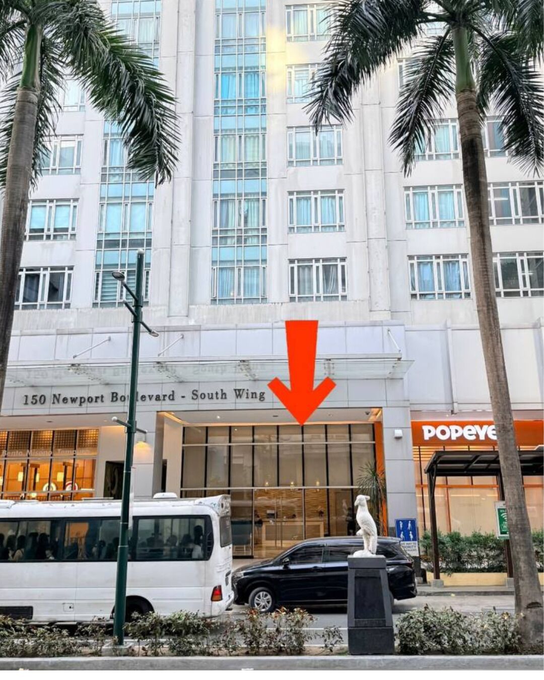 Un edificio a più piani con un ingresso ben visibile, un autobus parcheggiato di fronte e una statua in primo piano.