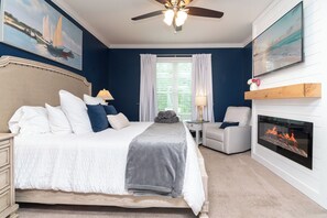 A bedroom with a bed, a fireplace, a ceiling fan, and artwork on the walls.