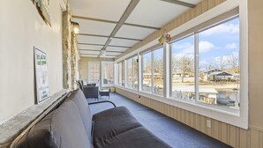 Een overdekt terras met een bank, een tafel en stoelen, grote ramen met uitzicht op de omgeving, en een muur met een poster.