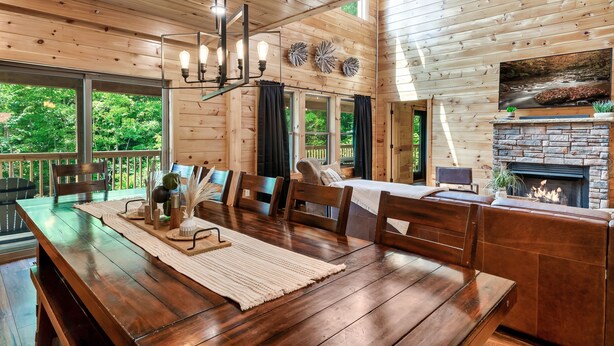 L’intérieur d’un chalet en bois comprend une table à manger, des chaises, une cheminée et une grande fenêtre donnant sur un paysage verdoyant.
