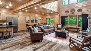 Une cabane spacieuse aux murs et au plancher en bois, comprenant un coin cuisine, un salon avec un canapé et des fauteuils, et de grandes fenêtres offrant une vue sur l’extérieur.