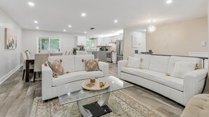Un salón moderno con sofás blancos, una mesa de centro de cristal y una cocina al fondo.