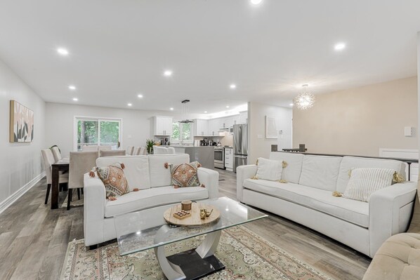 Un salón moderno con sofás blancos, una mesa de centro de cristal y una cocina al fondo.