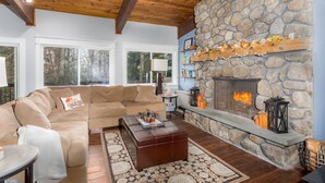 Una sala de estar acogedora con chimenea de piedra, vigas de madera en el techo y un amplio ventanal con vistas a un bosque.