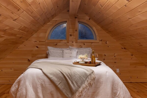Une chambre confortable située dans les combles, avec un plafond en bois, deux fenêtres de toit, un grand lit recouvert d’une couverture beige, des oreillers et un plateau sur lequel se trouvent une bouteille et des fleurs.