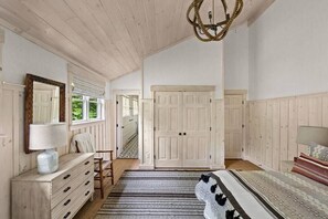 Un dormitorio con techo de madera, un candelabro, una cama con ropa de cama estampada, un aparador, un espejo y una puerta que da a otra habitación.