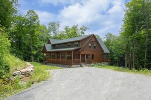 Une cabane en rondins avec un porche, entourée d’arbres et bordée d’une allée de gravier.