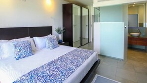 A modern hotel room with a large bed, a nightstand, a vase with flowers, and a view of the bathroom and kitchen through a glass door. - Beachfront Luxury with Incredible Ocean Views (Worthing)