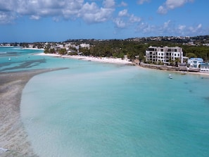 A coastal view with a beach, clear turquoise waters, and a building with multiple floors.