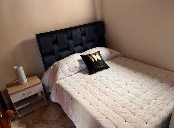A neatly made bed with a white quilt and a black pillow, a wooden nightstand with a white vase, and a tiled floor.