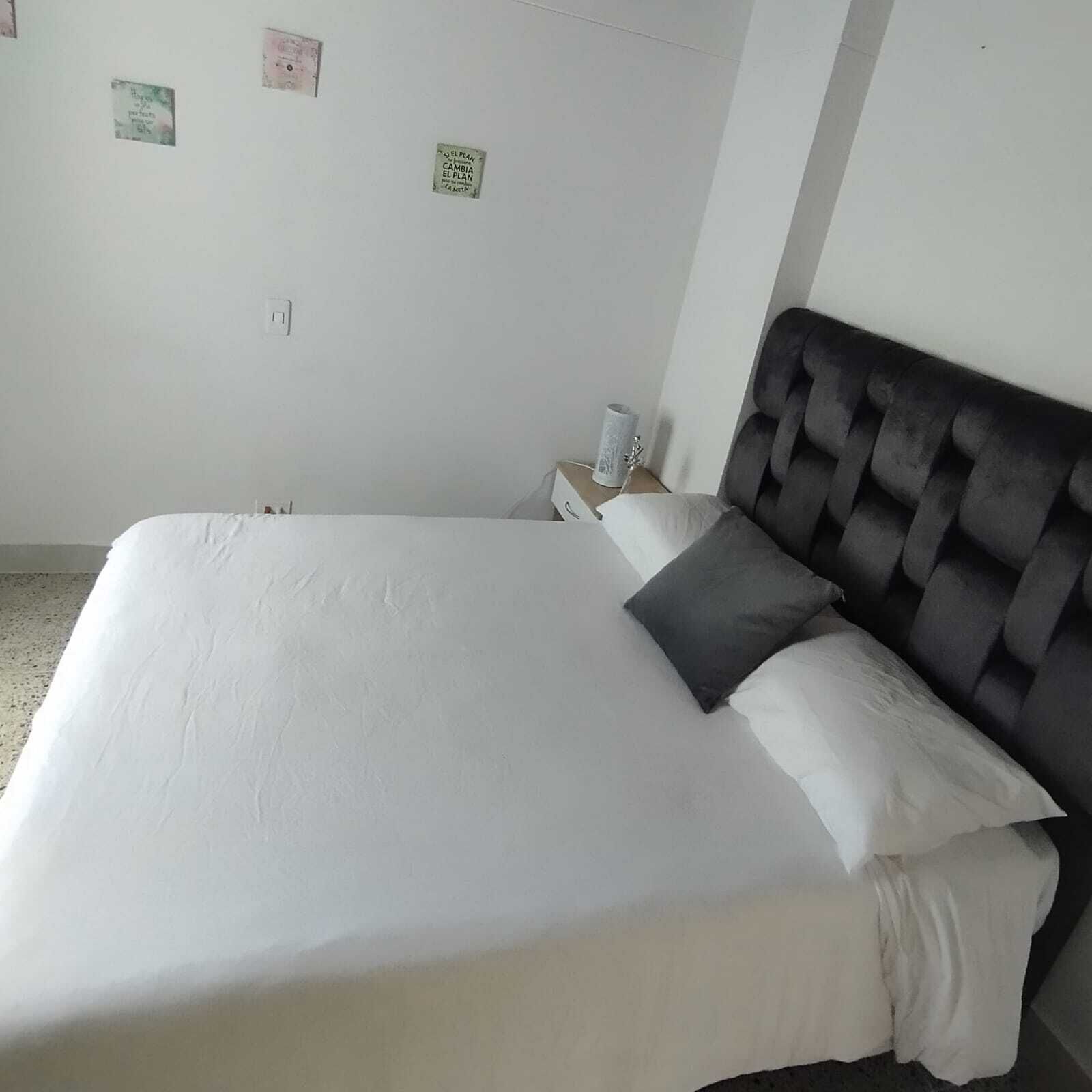 A neatly made bed with white linens and a dark headboard in a simple room with white walls.