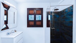 A bathroom with a white vanity, a large mirror, a shower with dark blue tiles, and a window with wooden shutters.