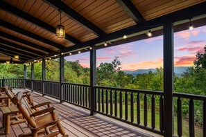 Un espacio de estar al aire libre cubierto con bancos de madera y barandilla, con vistas a un paisaje pintoresco con árboles y un atardecer.