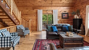 Ein rustikales Blockhaus-Interieur mit Treppe, karierten Sofas, einem Ledersofa, einem gemusterten Teppich und einem Vintage-Koffer.