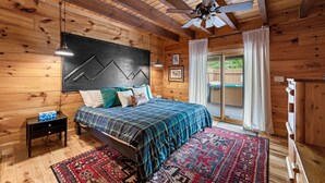 Ein Schlafzimmer einer Holzhütte mit einem Bett, einem Nachttisch, einem Deckenventilator und einem großen Fenster mit Vorhängen.