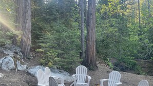 Un groupe de fauteuils Adirondack est disposé autour d’un foyer en pierre, dans une zone boisée où poussent de grands arbres.