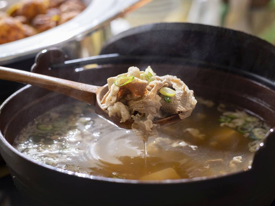 Primer plano de una sopa que se sirve con palillos, con una olla y una cuchara visibles al fondo.