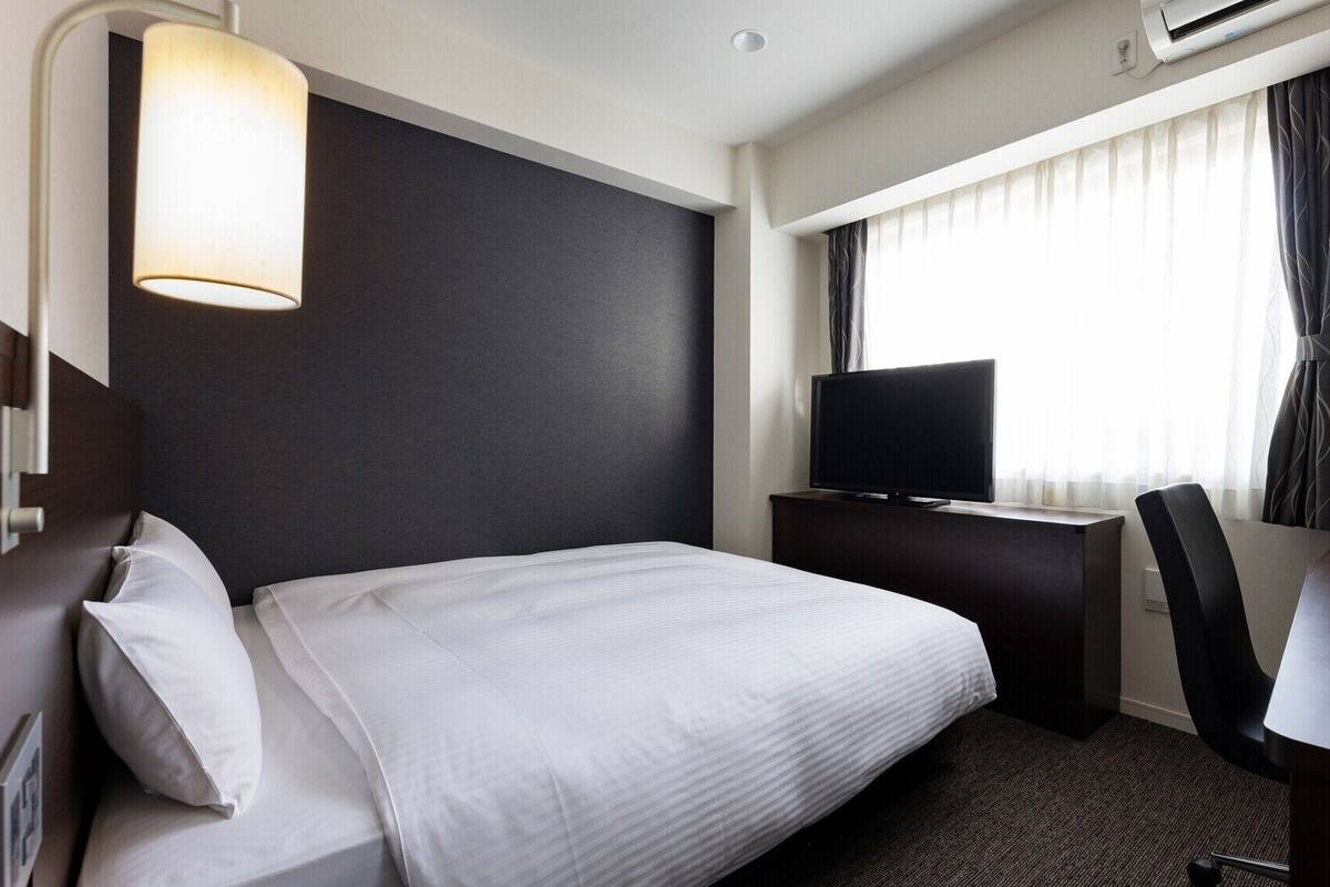 Una habitación de hotel con cama, escritorio, televisión y ventana con cortinas.
