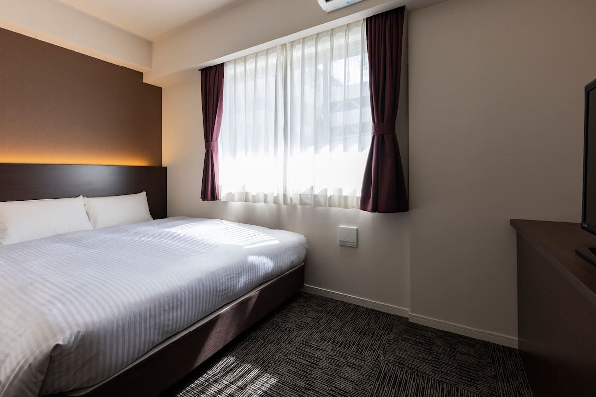 Habitación de hotel con una cama grande, un equipo de aire acondicionado, una ventana con cortinas y un termostato instalado en la pared.