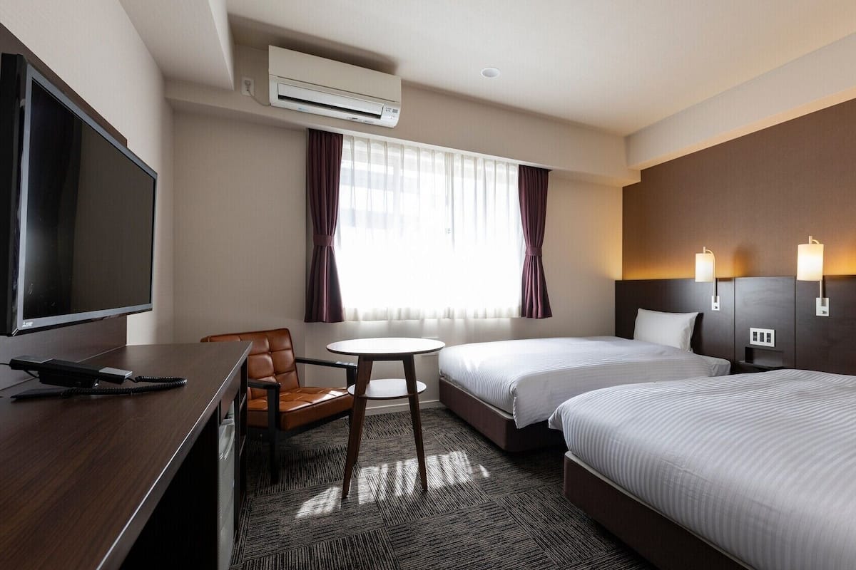 Habitación de hotel con escritorio, dos camas, televisor, una mesita y ventana con cortinas.