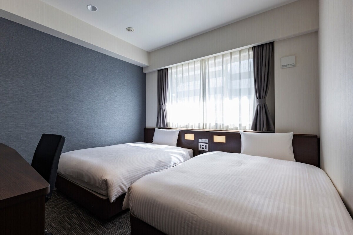 Habitación de hotel con dos camas, un escritorio y un amplio ventanal con cortinas.