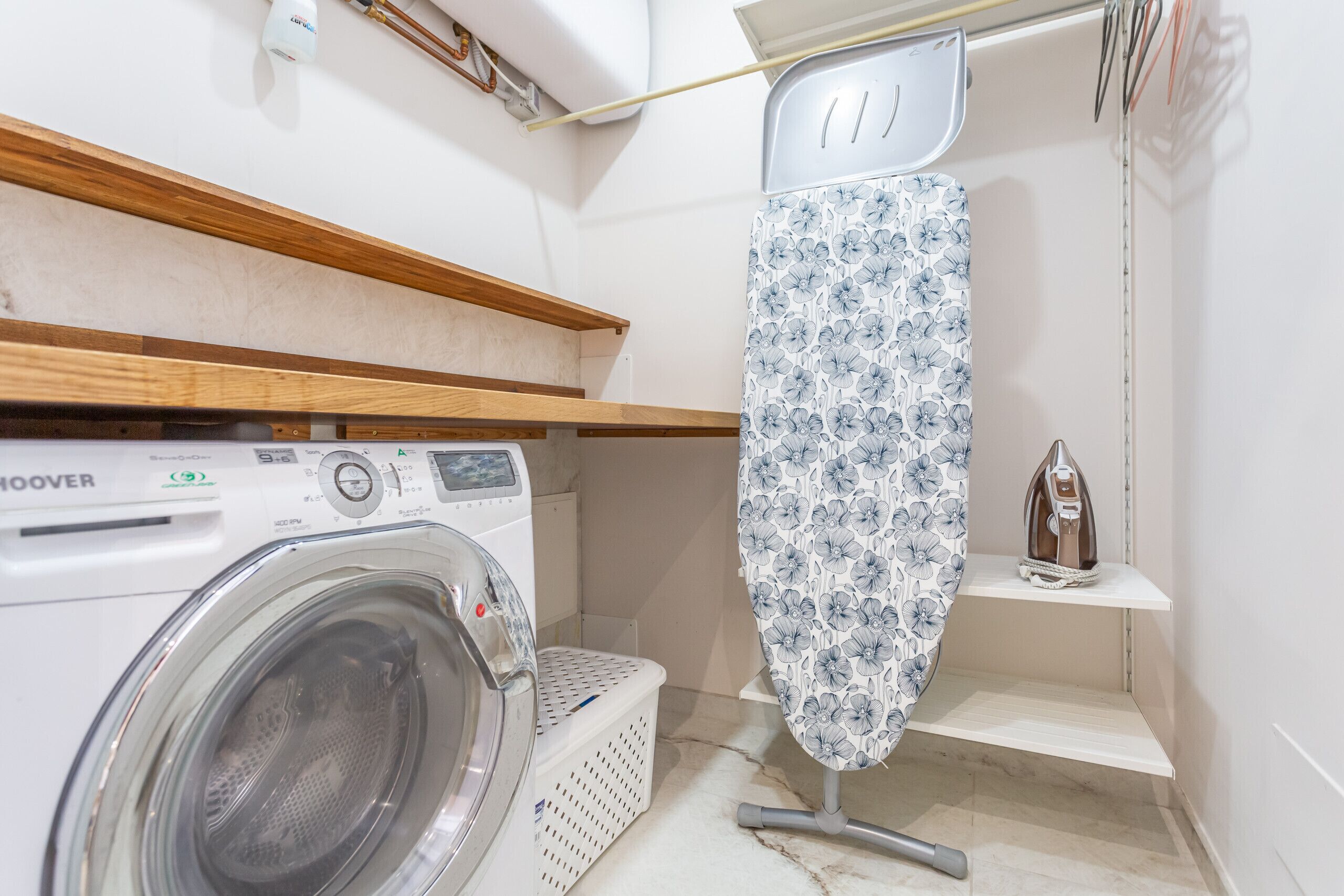 A compact laundry area with a front-loading washing machine, an ironing board, and a small shelf with a kettle.