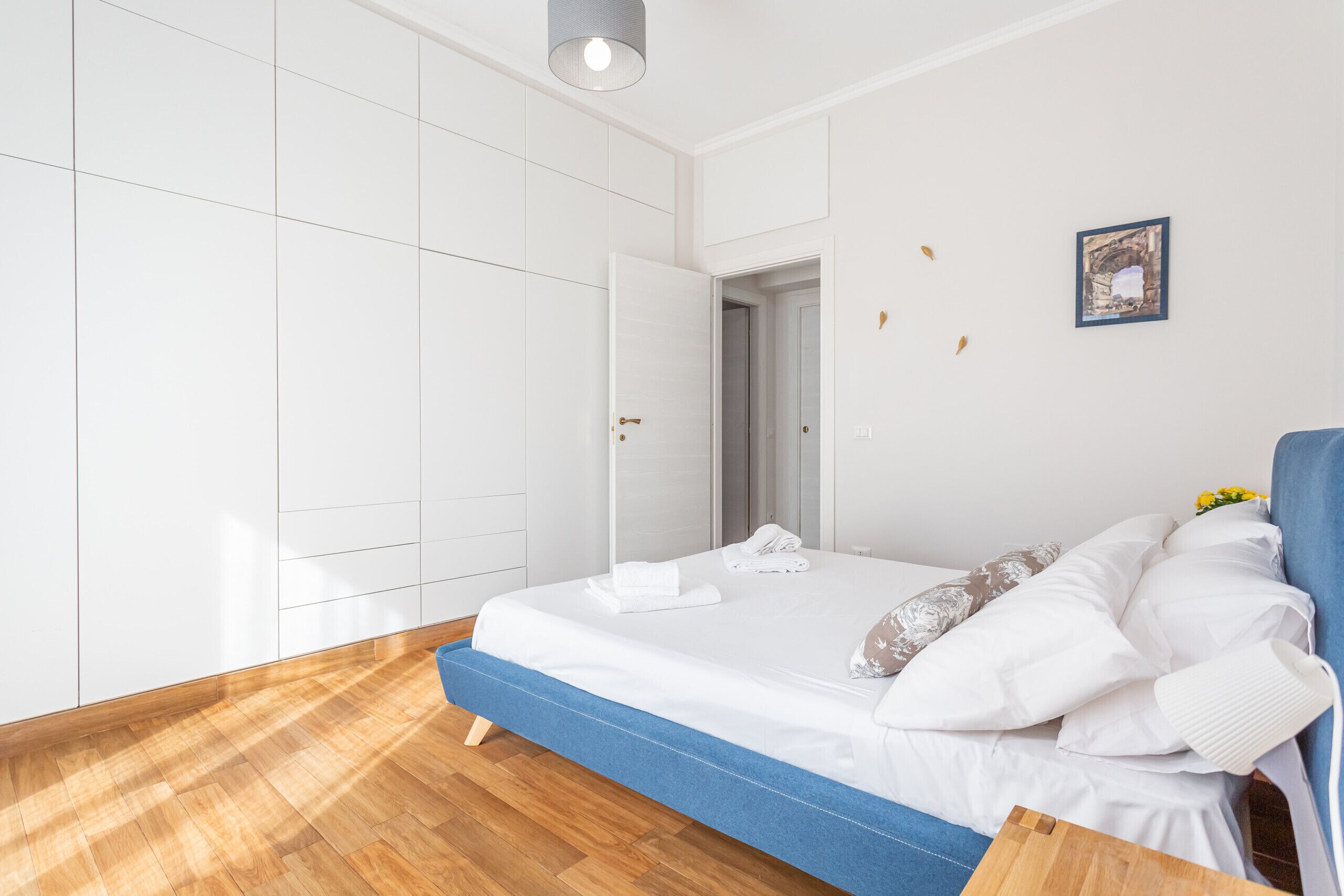 A modern bedroom with a large bed, white walls, and a wooden floor.